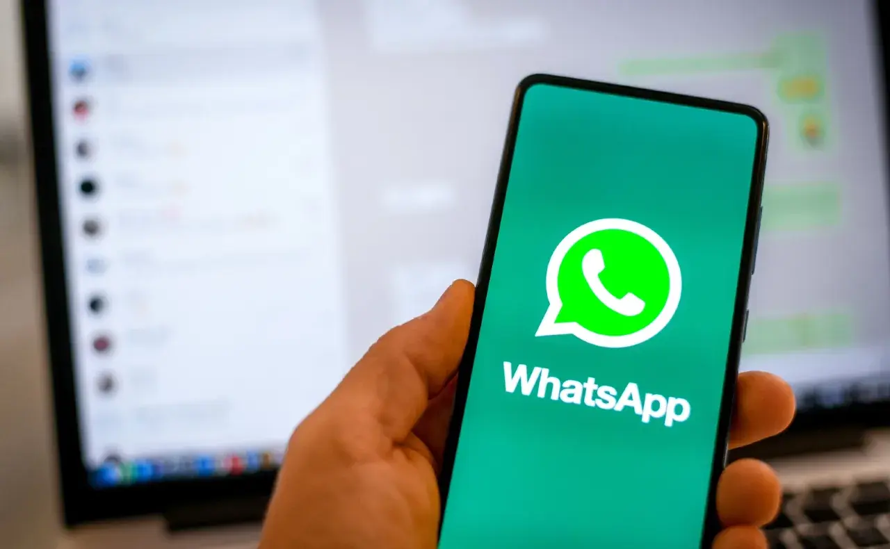 Dłoń trzyma smartfon z logo WhatsApp. W tle widać ekran laptopa z otwartym programem do komunikacji, sugerującym użycie WhatsApp na komputer.