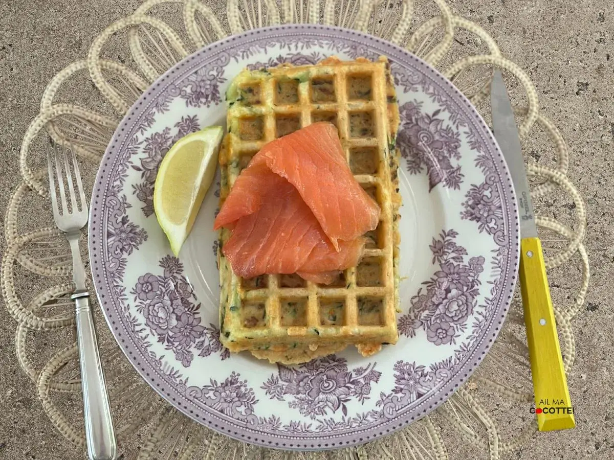 Gaufres salées garnies de saumon fumé et d'un quartier de citron, sur une assiette ornée, avec une fourchette et un couteau à manche jaune.