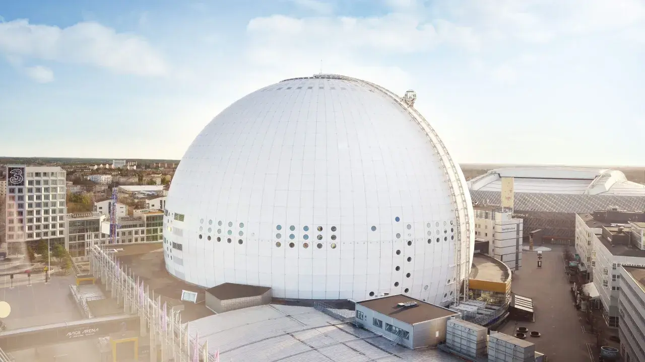 En solig dag vid Globen, Stockholm. Den ikoniska sfäriska arenan dominerar landskapet, omgiven av moderna byggnader och en klarblå himmel.