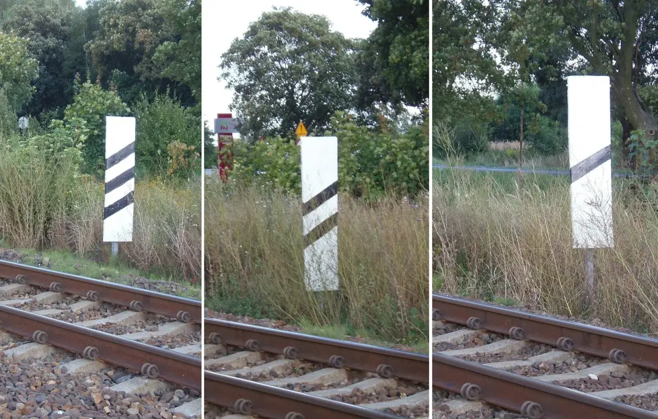 Trzy białe słupki z czarnymi pasami, wskaźnik w11a, stoją przy torach kolejowych.