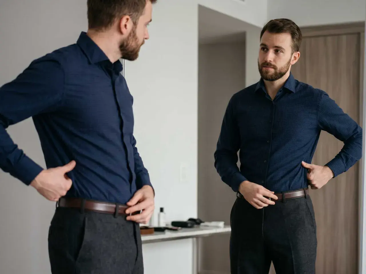Mann im Spiegel prüft verschiedene Arten von Hemden, trägt ein dunkelblaues Hemd und graue Hose.