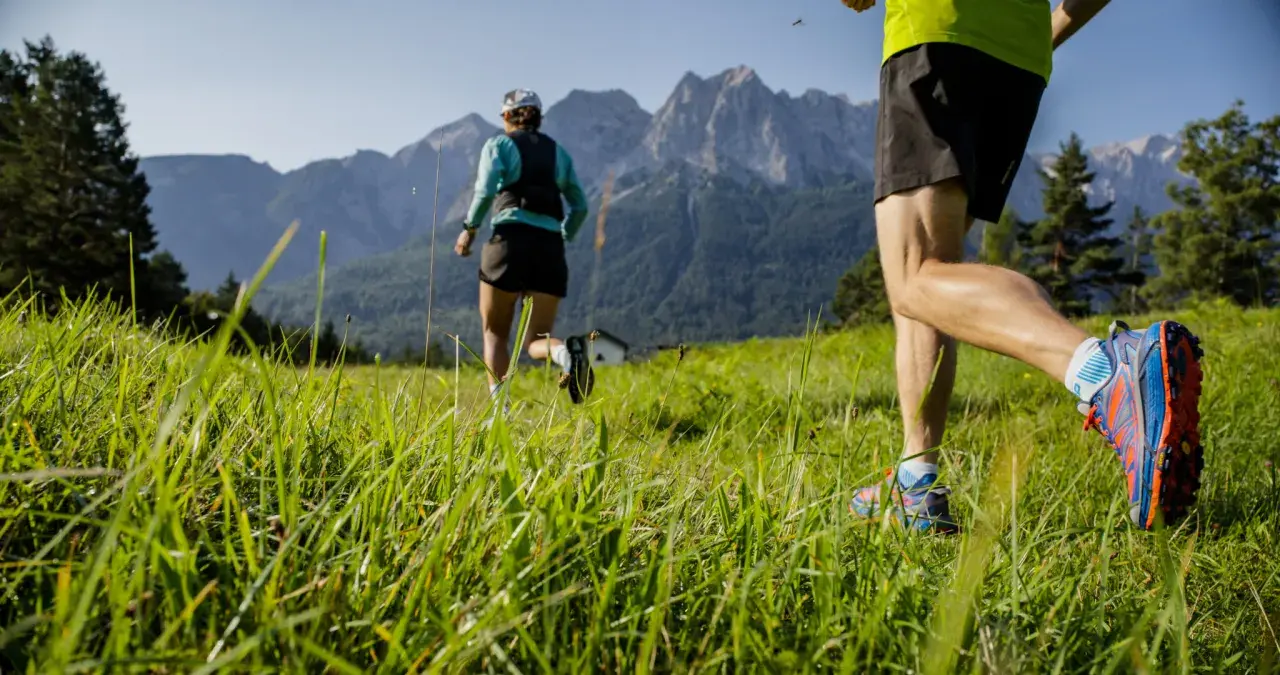 Zwei Läufer genießen das Zugspitze Laufen auf einer grünen Wiese vor majestätischen Bergen.