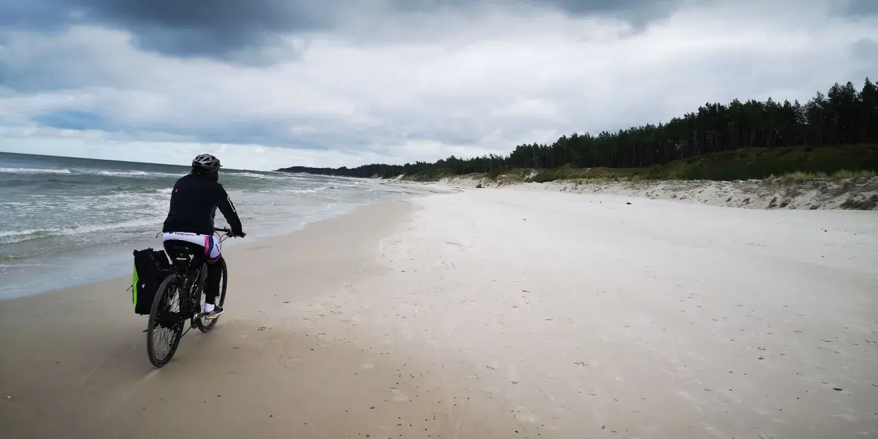 Rowerzystka przemierza piaszczystą plażę na mierzei sarbskiej, z widokiem na wzburzone morze i sosnowy las.