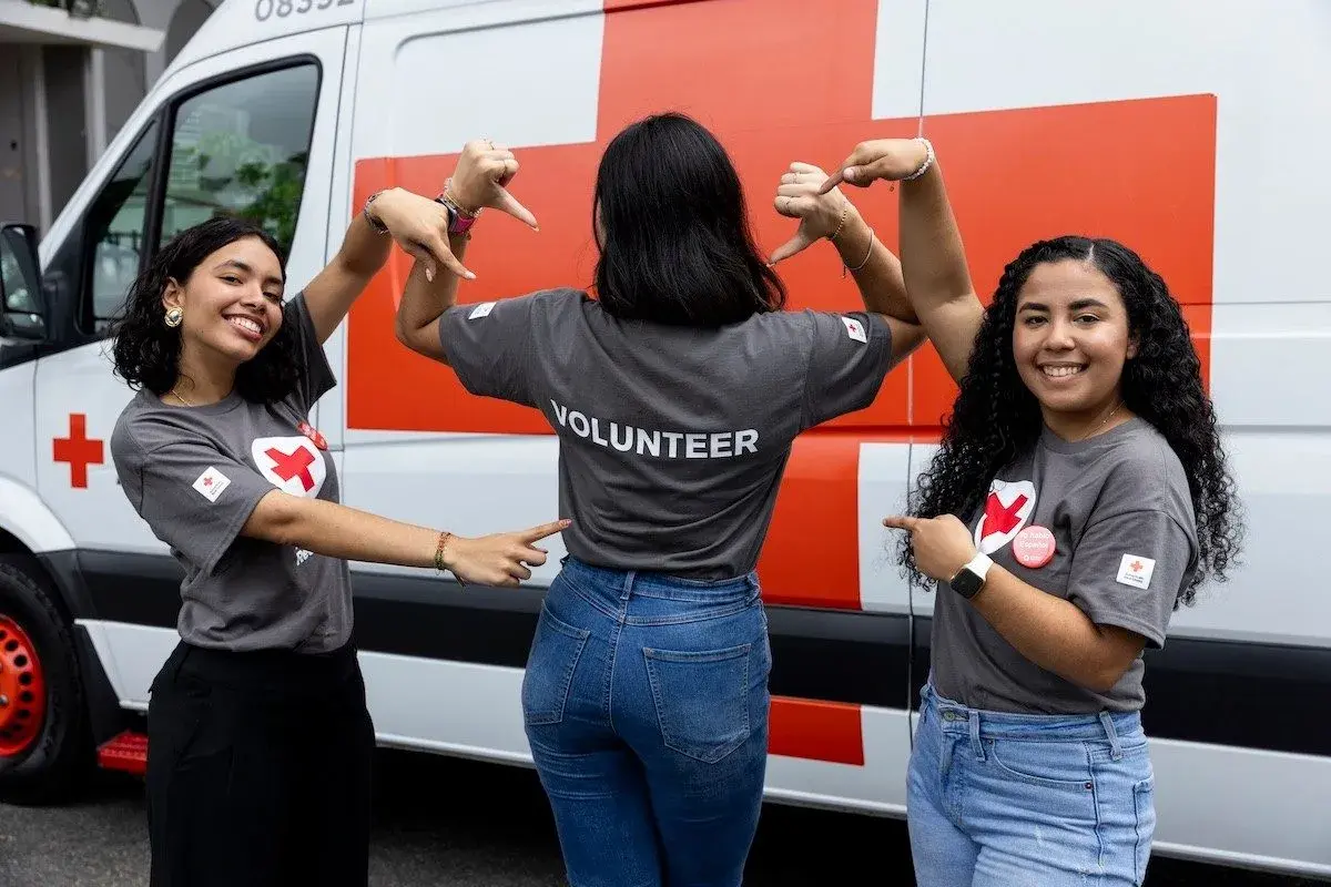 Three young women in "VOLUNTEER" shirts pose in front of a Red Cross van, smiling and pointing.