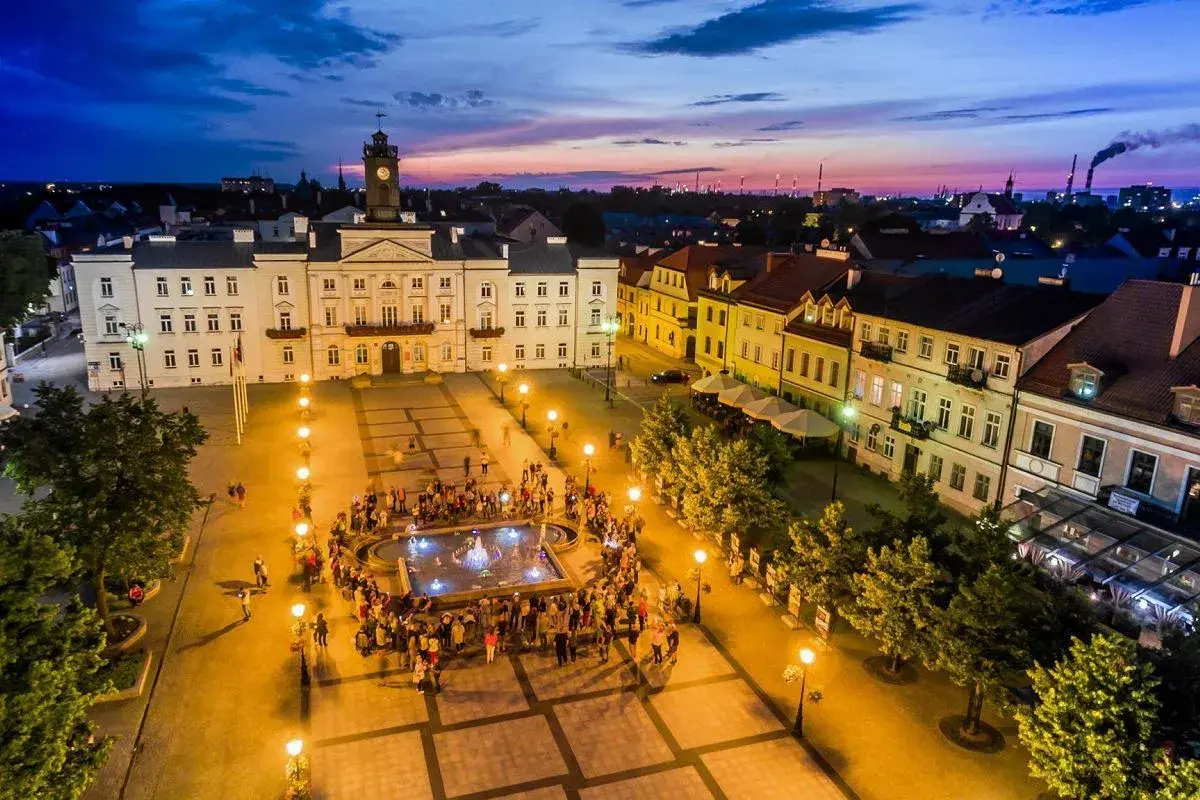 Wieczorny stary rynek płock, z ratuszem i fontanną, oświetlony latarniami, z tłumem ludzi.