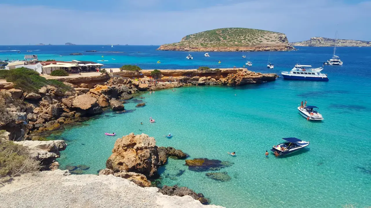 Plage idyllique à Ibiza avec eaux turquoise, rochers, bateaux et baigneurs. Parfait pour savoir où aller à Ibiza.