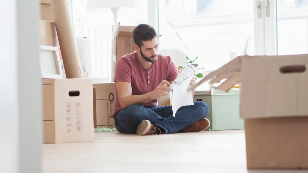 Hombre revisa un plano rodeado de cajas, preparándose para la mudanza. Podría ser el **contrato compra venta** de su nuevo hogar.