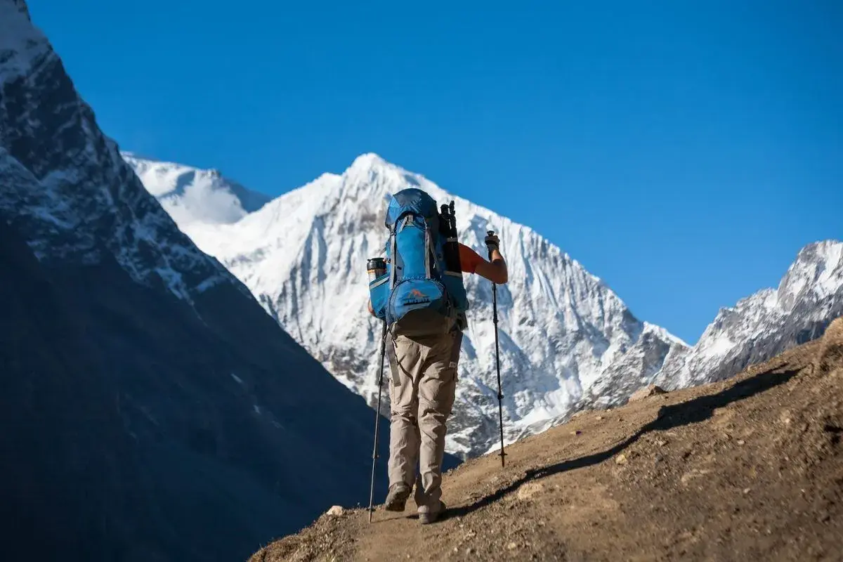 Un excursionista con mochila azul asciende un sendero polvoriento, bastones en mano, ante majestuosas montañas nevadas. Una aventura de trekking Manaslu.