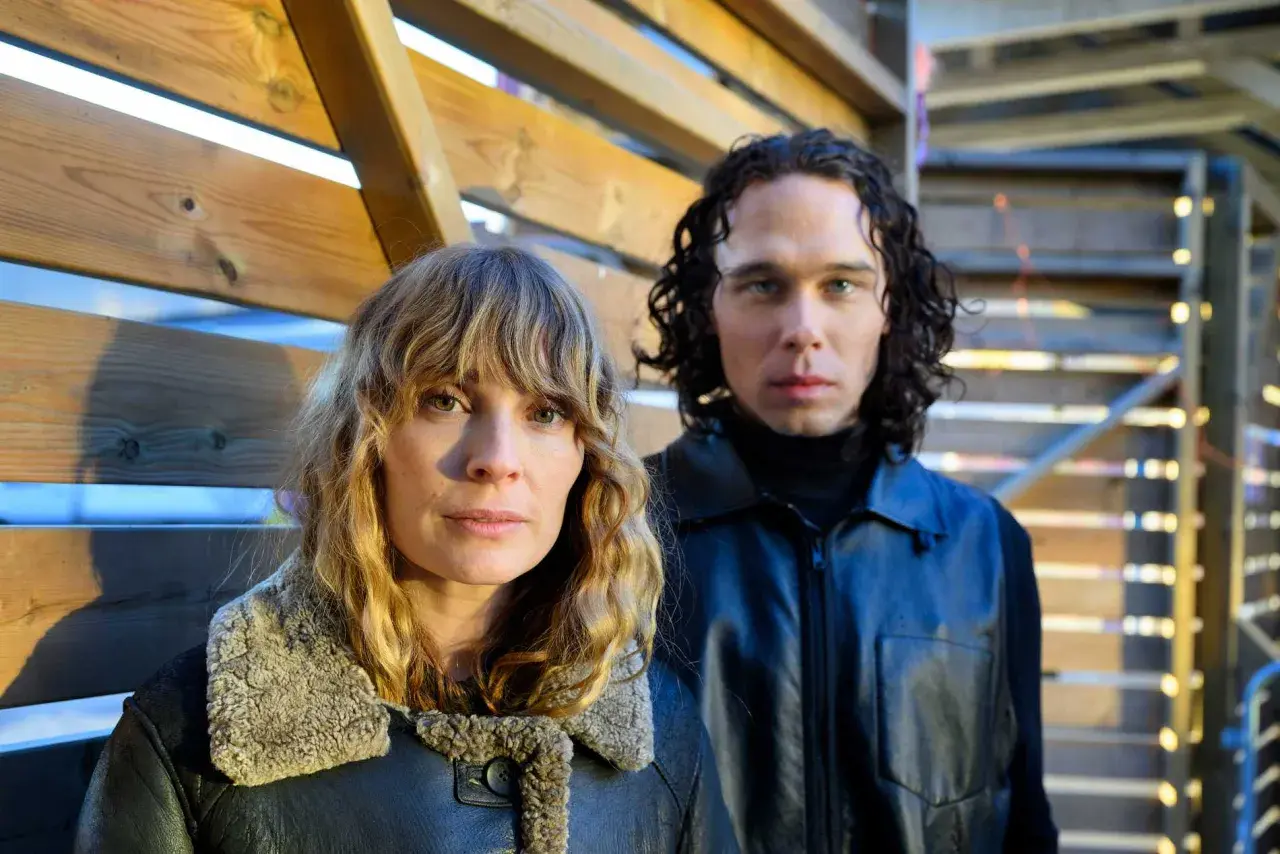 Två personer, en kvinna med blont hår och en man med lockigt mörkt hår, står framför en träkonstruktion. De är skådespelare från rollistan i Knutby (tv-serie).