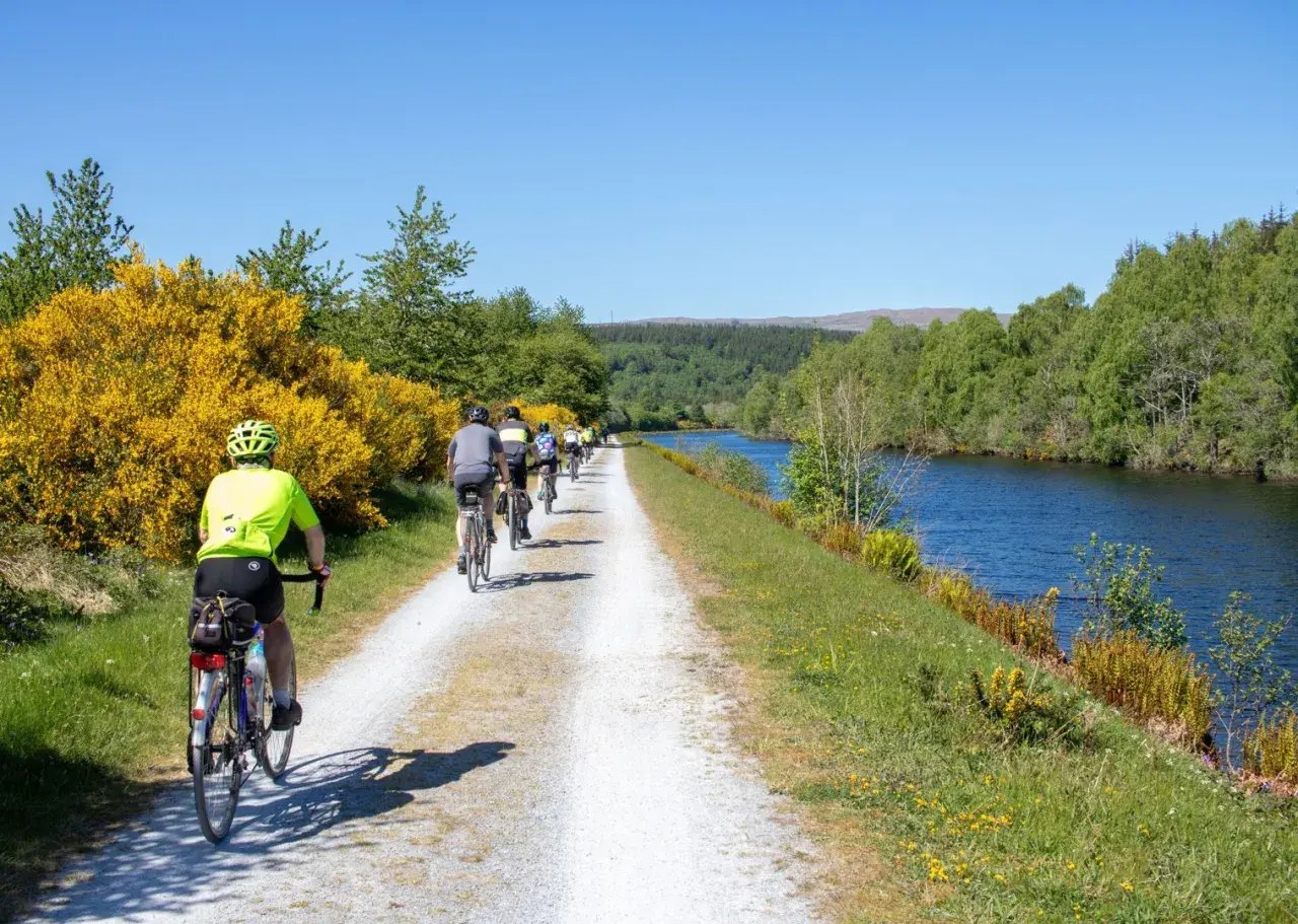 A group enjoys cycling in Scotland along a canal path, with bright yellow gorse bushes and lush green trees lining the route.