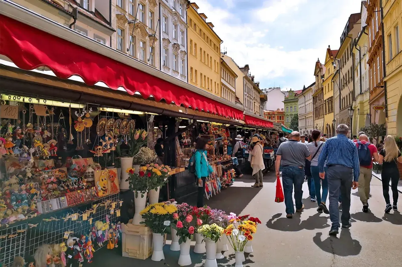 En livlig marknad i Prag med stånd fyllda av leksaker och blommor. Folk promenerar längs den soliga gatan.