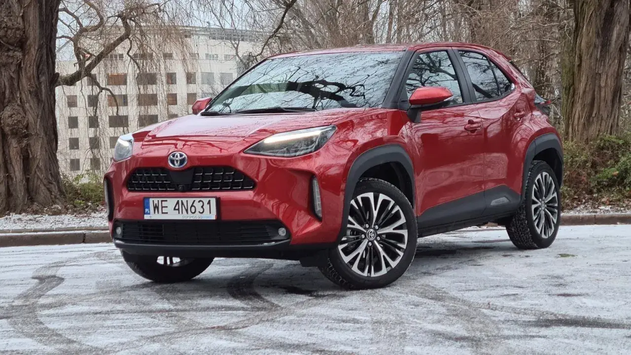 Czerwony Toyota Cross Hybrid na tle drzew i budynku. Auto stoi na ośnieżonej nawierzchni.