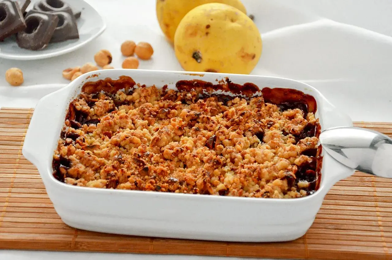 Delizioso crumble pere e cioccolato appena sfornato, con pere mature e nocciole sullo sfondo.