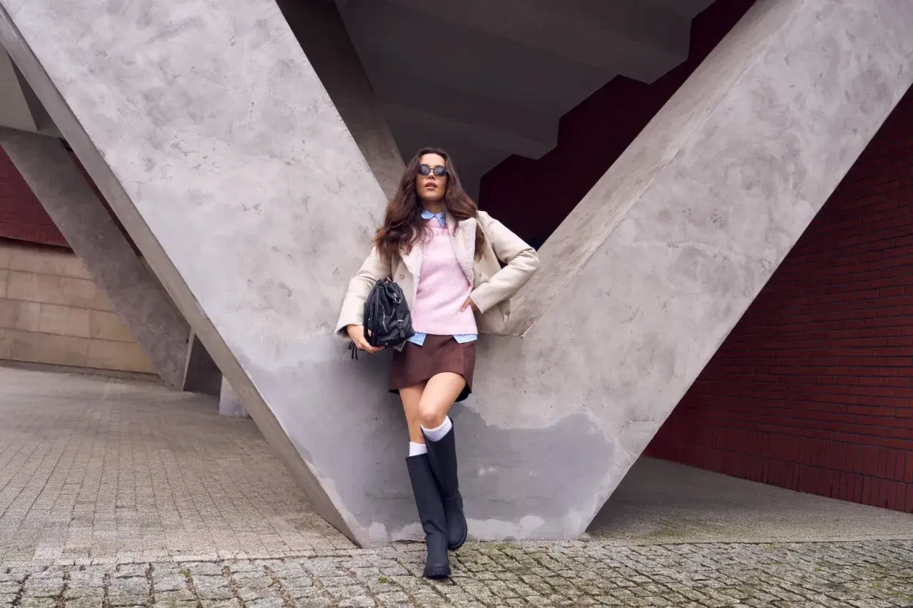 Dziewczyna w różowym sweterku do brązowej spódniczki, beżowej kurtce i czarnych kozakach pozuje na tle betonowej konstrukcji.