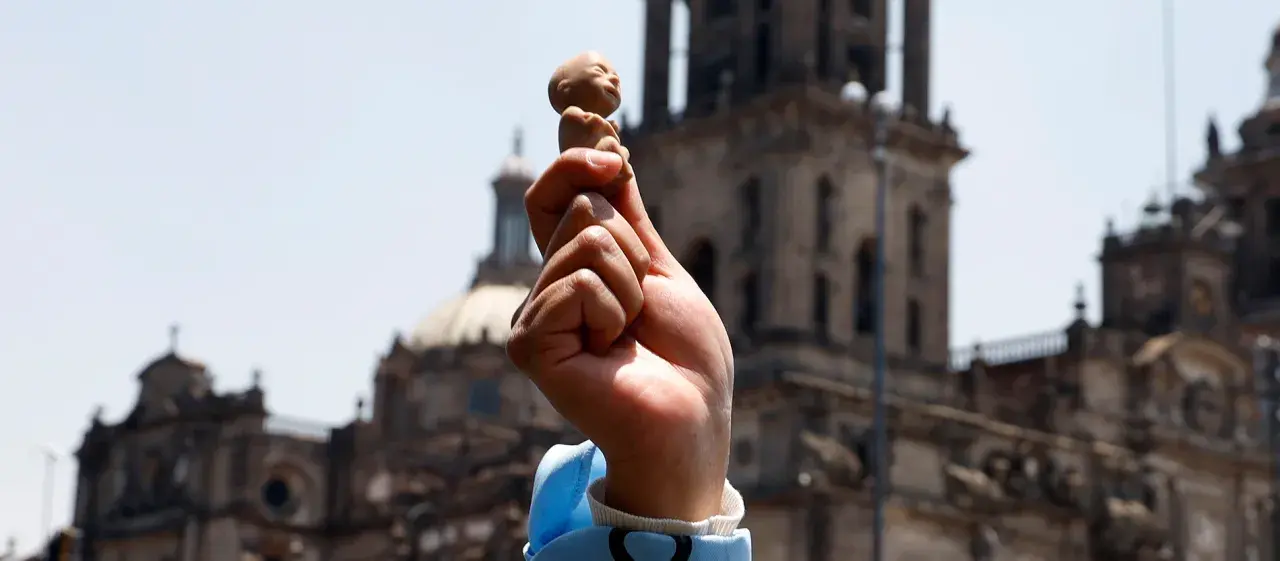 Hand hält Fötus-Figur vor Kirche. Debatte um evangelische Kirche und Abtreibung.