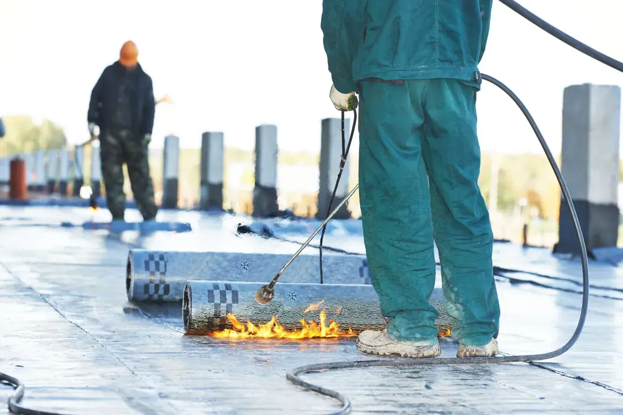 Arbeiter schweißen Bitumenbahnen, um ein Flachdach abzudichten. Flammen erhitzen das Material für eine sichere Verbindung.