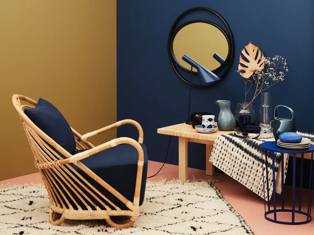 Ein Rattan-Sessel mit dunkelblauem Kissen steht auf einem flauschigen Teppich. Welche Farbe passt zu Gold? Hier: ein Spiegel mit goldenem Rahmen und ein gelber Wandbereich.