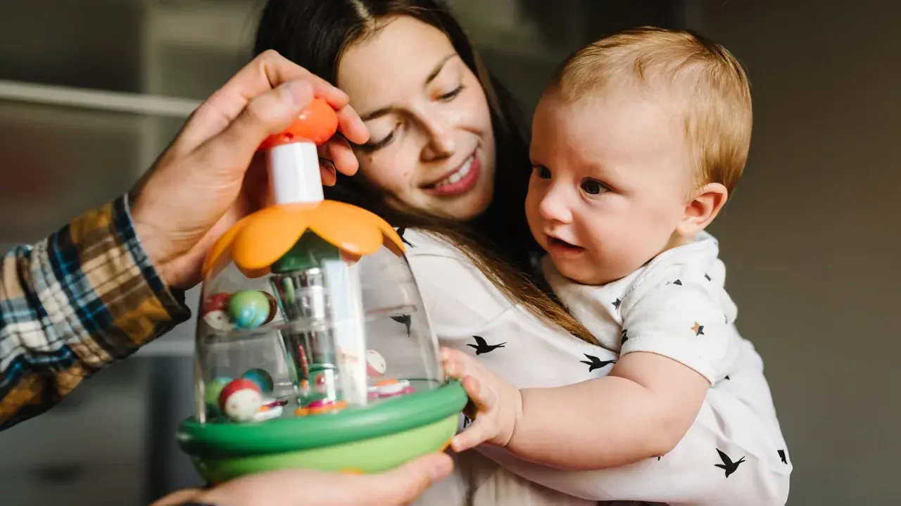 Mama i tata pokazują dziecku zabawkę. Maluch, choć nie jest jeszcze dwulatkiem, z zaciekawieniem kręci głową, obserwując kolorowe kulki.