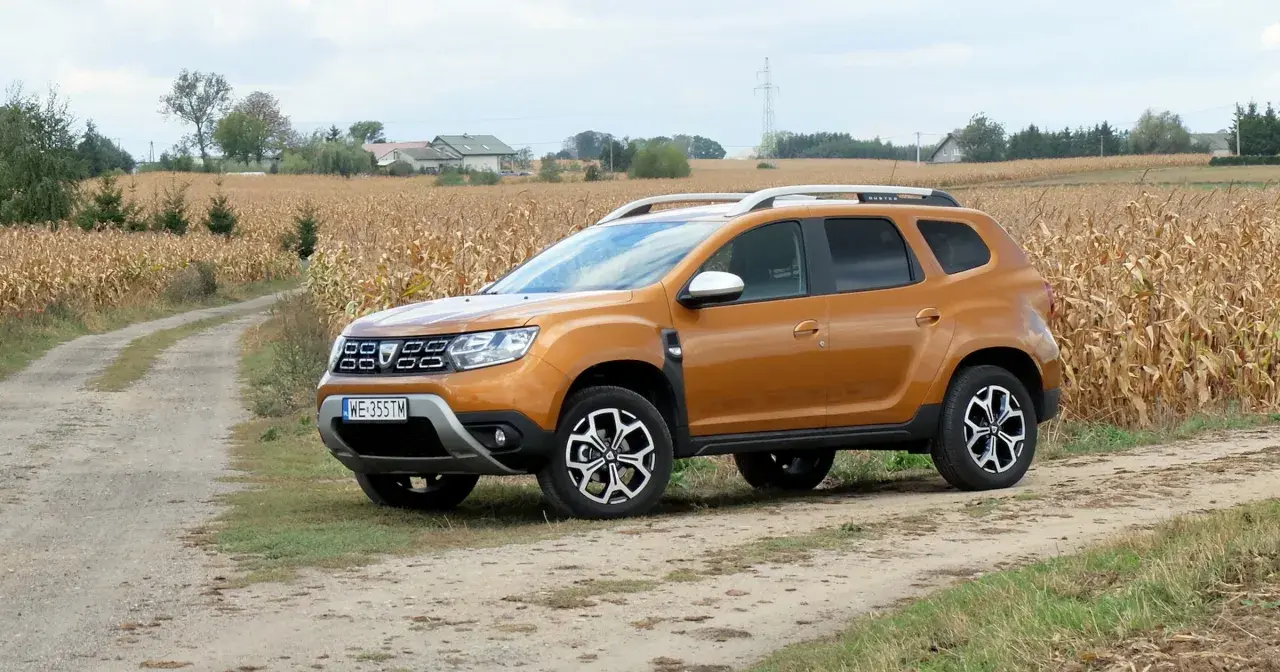 Pomarańczowy Dacia Duster 1.6 benzyna na polnej drodze, w tle pole kukurydzy i domy.