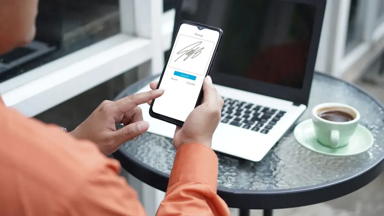 Persona usando su móvil para firmar online documento. En la mesa hay un portátil y una taza de café.