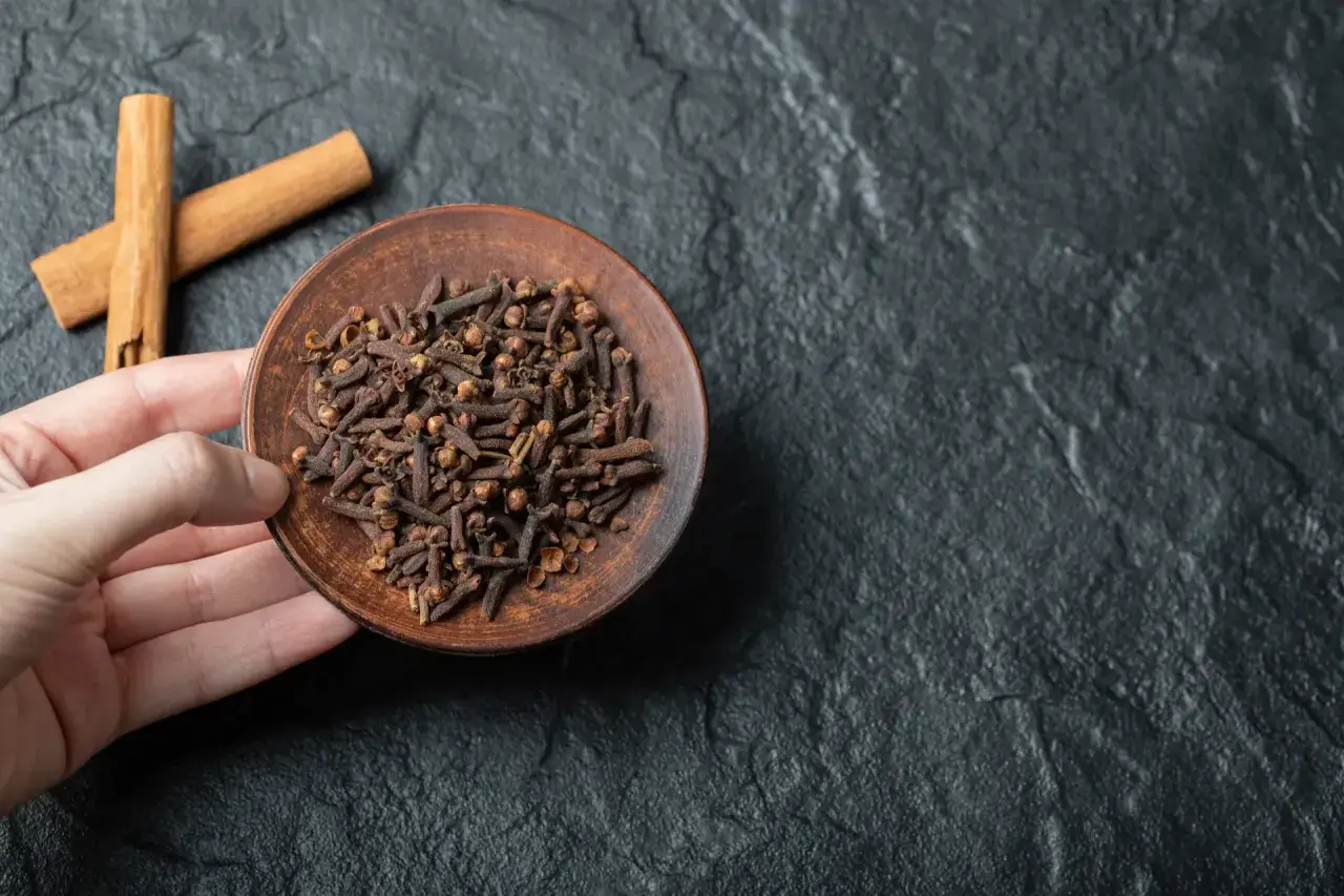 Un plato de clavo de olor, un remedio natural para el dolor de muelas, junto a dos ramas de canela sobre una superficie oscura.