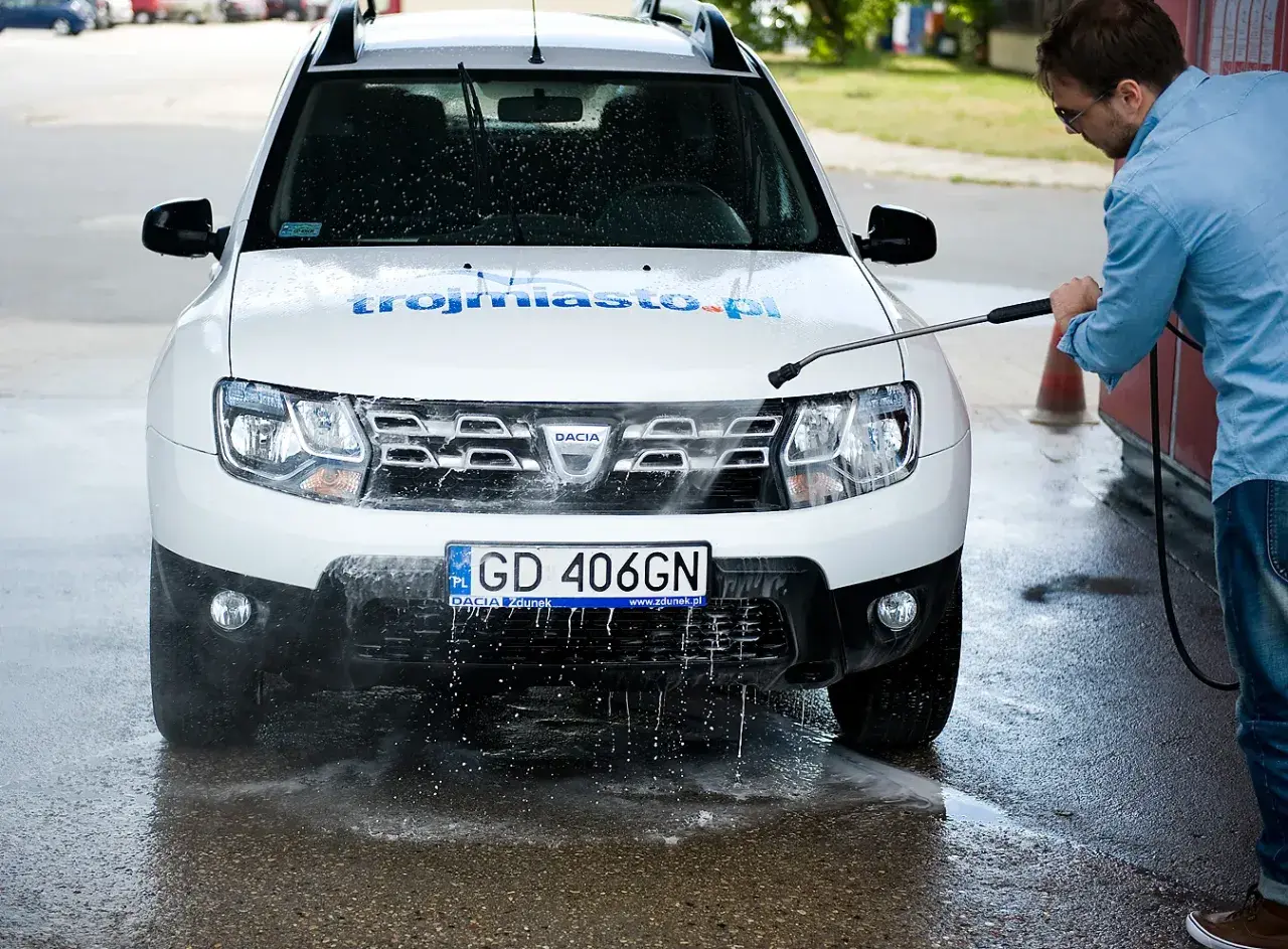 Mężczyzna myje białe Dacia Duster na myjni bezdotykowej, spryskując przód auta wodą pod ciśnieniem.