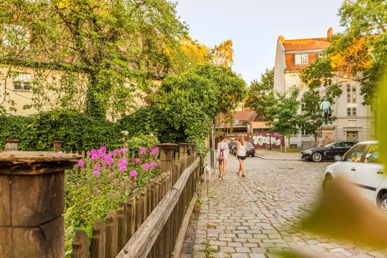 Zwei Frauen spazieren mit Hund über Kopfsteinpflaster in Rixdorf, Berlin. Bunte Blumen und üppiges Grün säumen den Weg.