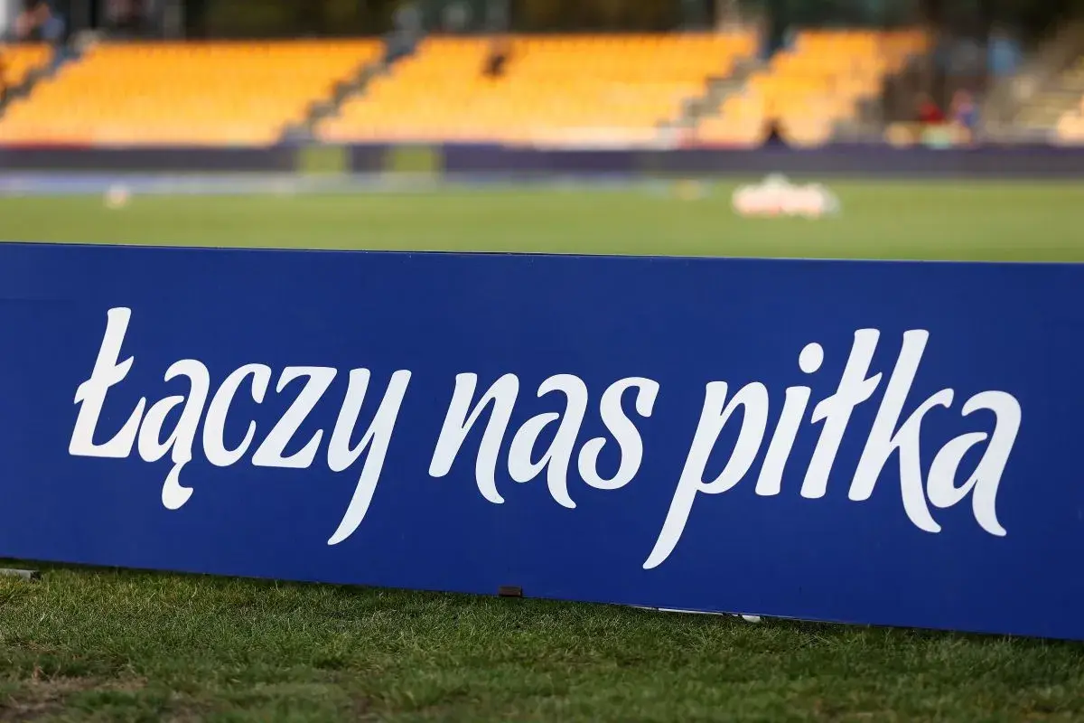 Niebieska tablica z napisem "łączy nas piłka" na tle stadionu. To motto, które jednoczy kibiców i prezesa PZPN.