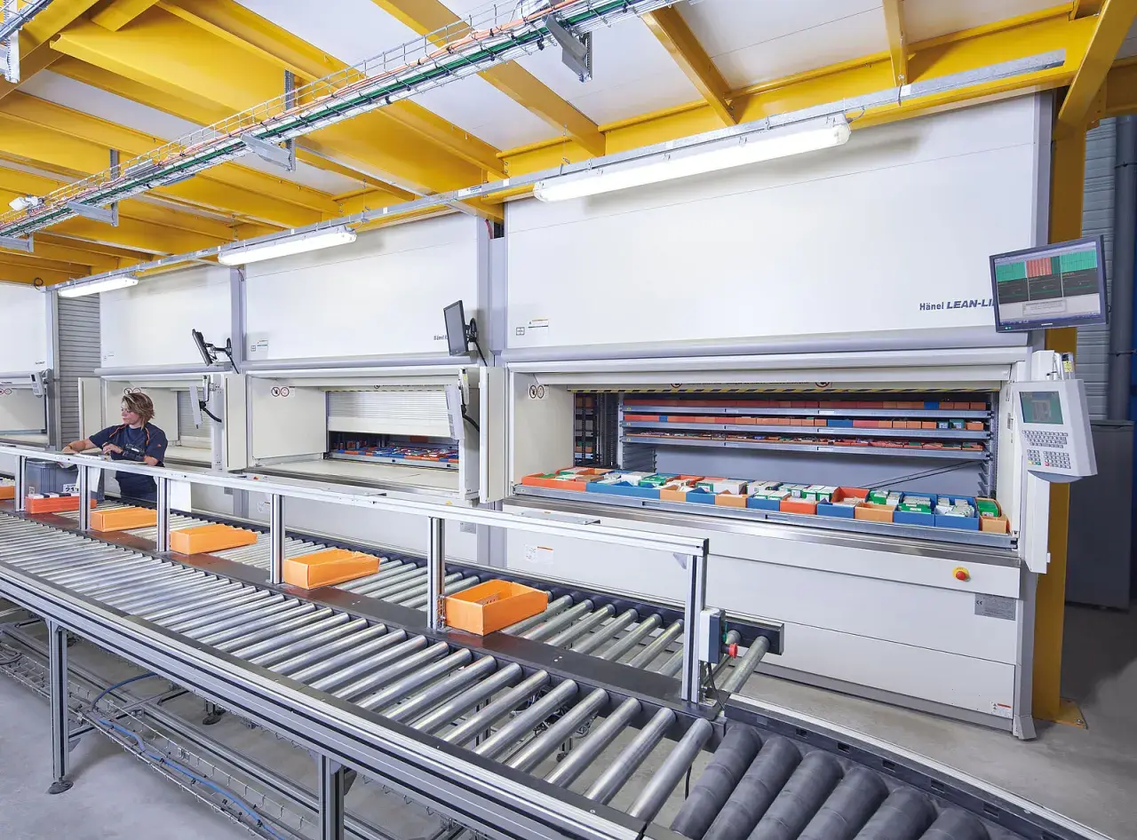 Ein Kommissionierlager mit automatisierten Lagersystemen. Eine Mitarbeiterin entnimmt Teile, während Behälter auf einem Rollenband transportiert werden.