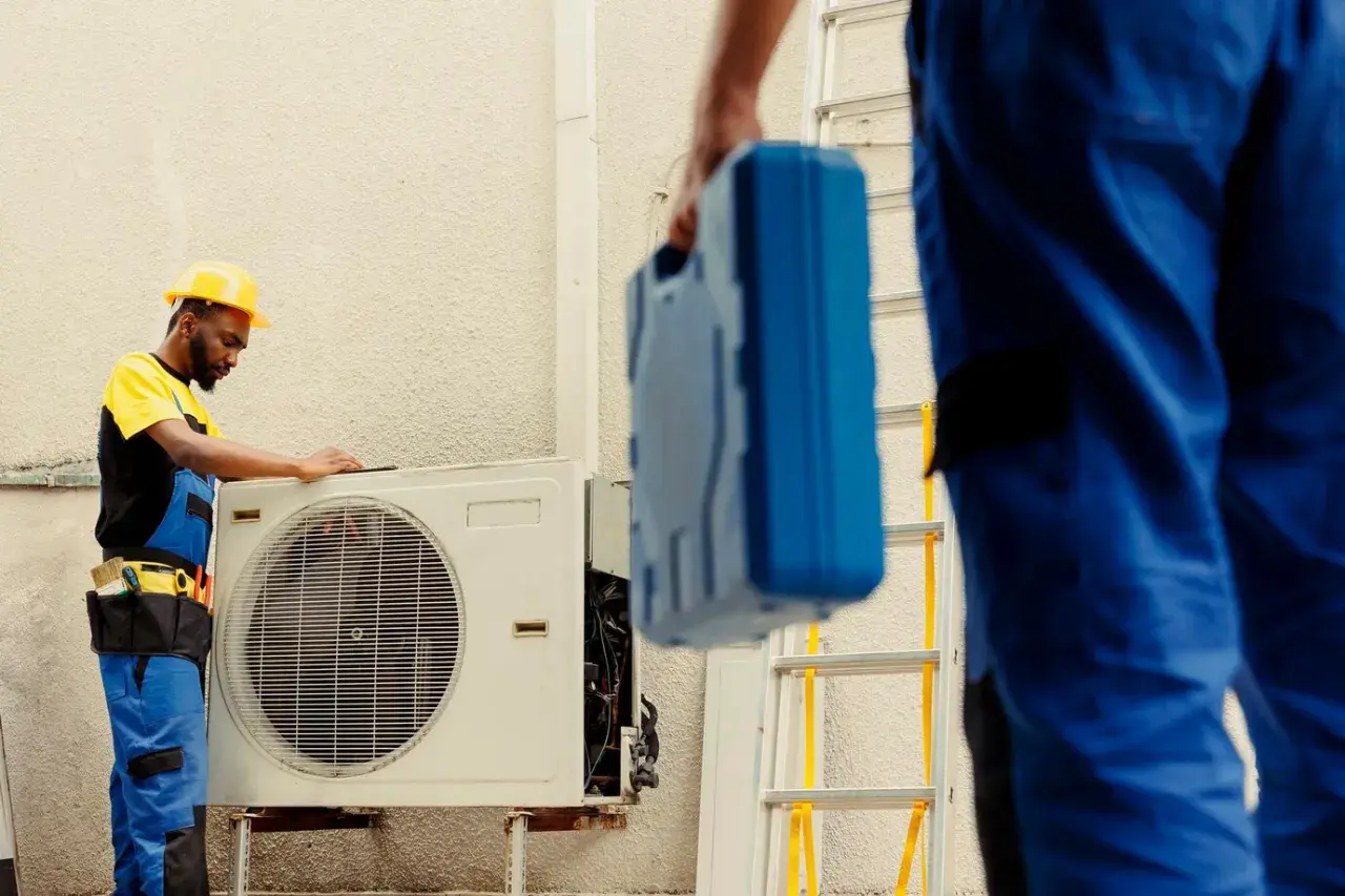 Técnicos revisan unidad exterior de aire acondicionado que dejó de funcionar de repente. Uno con casco amarillo examina la máquina, otro lleva una caja de herramientas azul.