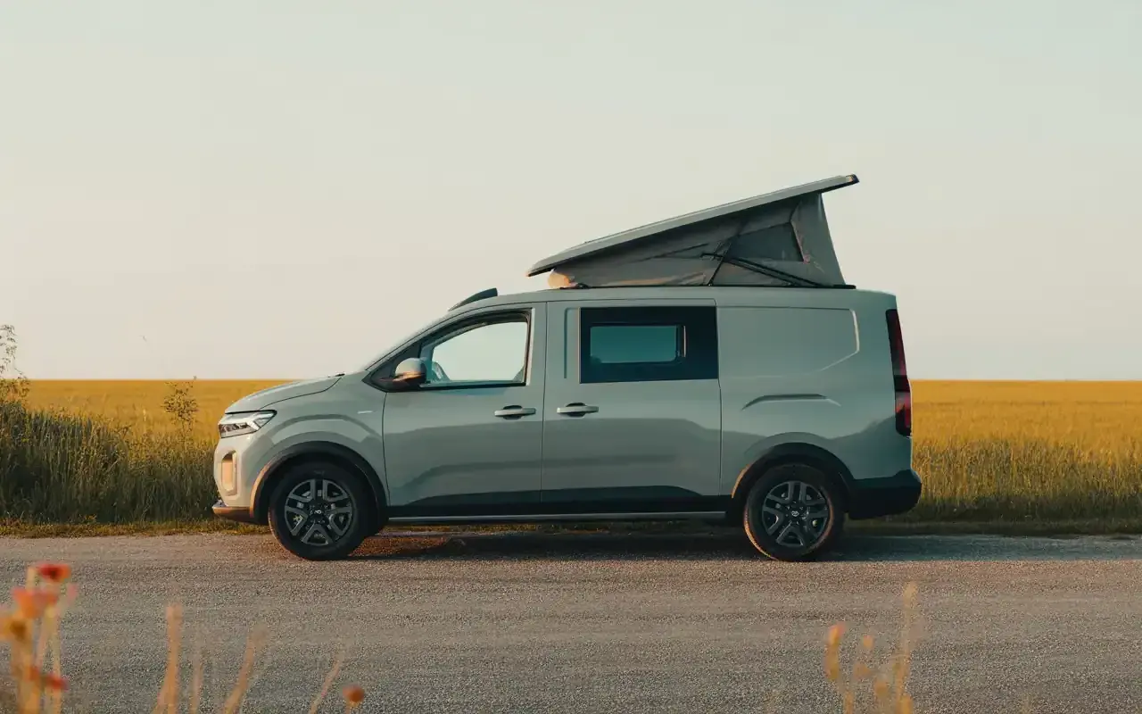 Un Dacia Sandman gris, prêt pour l'aventure, avec son toit relevable ouvert, stationné au bord d'un champ doré au coucher du soleil.