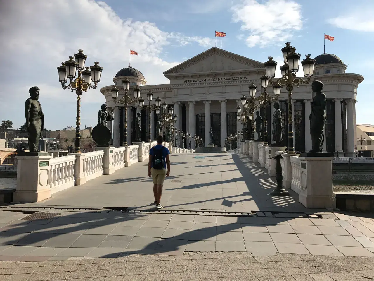 Un hombre camina por un puente adornado con estatuas y farolas, frente al Museo Arqueológico de Macedonia. ¡Un lugar imperdible que ver en Skopje!