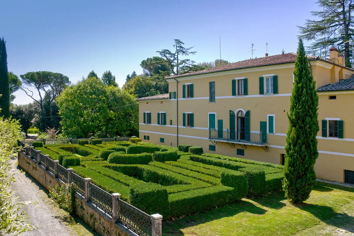 Villa Gracciano della Seta: un giallo edificio con un giardino geometrico di siepi verdi e un cipresso slanciato.