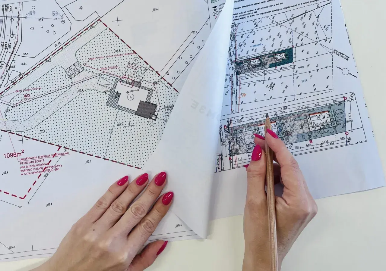 Rysunek techniczny z naniesionym projektem przyłącza wodociągowego. Dłoń z paznokciami malowanymi na różowo trzyma ołówek nad dokumentem.