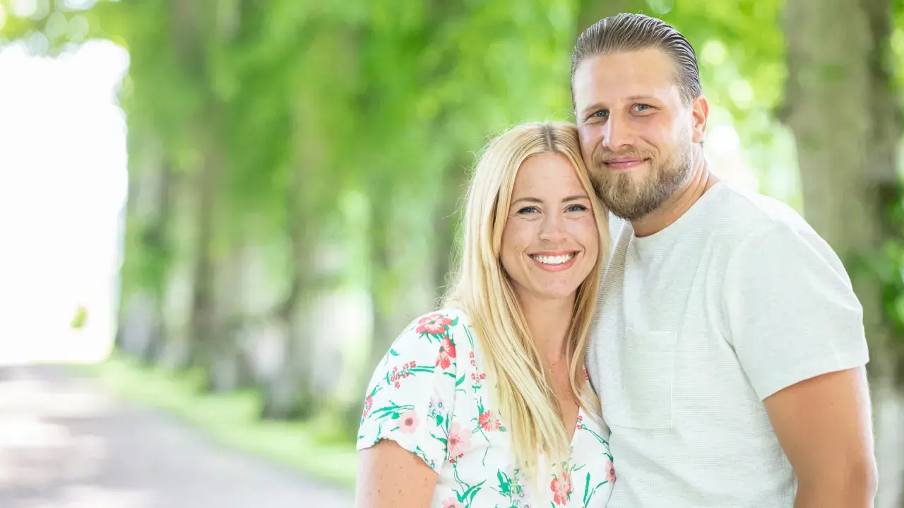 Ett leende par, Nanna Olasdotter Hallberg och hennes partner, njuter av en sommardag utomhus bland gröna träd.