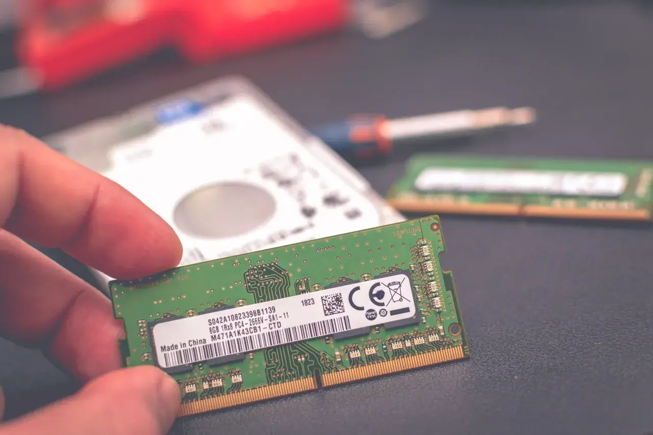 Dłoń trzyma moduł pamięci RAM 8 GB. Obok leży dysk twardy i drugi moduł RAM, sugerując wybór: 8 czy 16 GB RAM w laptopie.