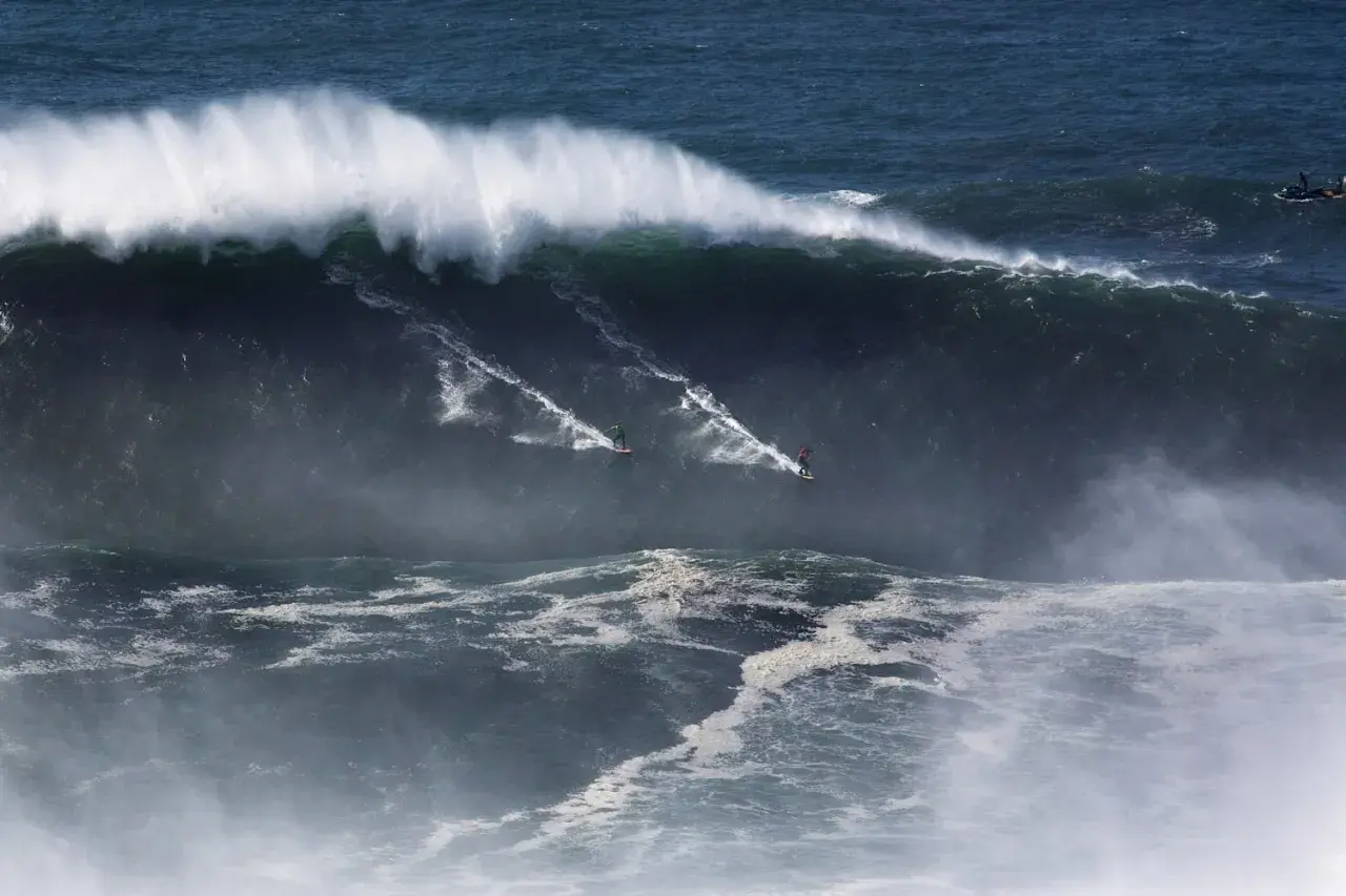 Surferzy ścigają się po gigantycznych nazare falach, tworząc białe smugi na ciemnym oceanie.