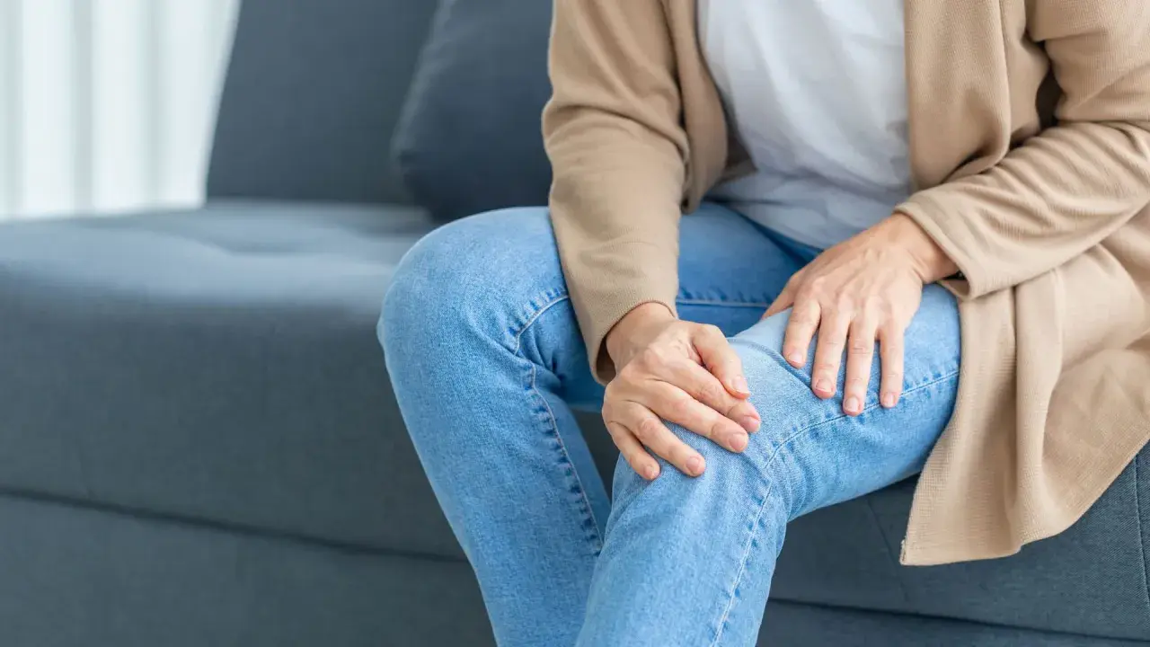 Frau sitzt auf Sofa, hält ihr Knie. Sie spürt ein unangenehmes Kribbeln in den Beinen.