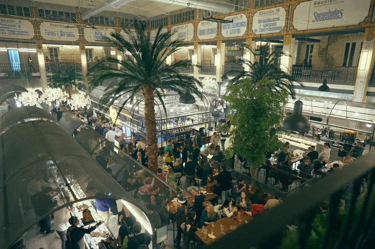 Un bullicioso mercado interior con palmeras, gente comiendo y comprando. El ambiente recuerda a un mercado de la imprenta, lleno de vida y actividad.