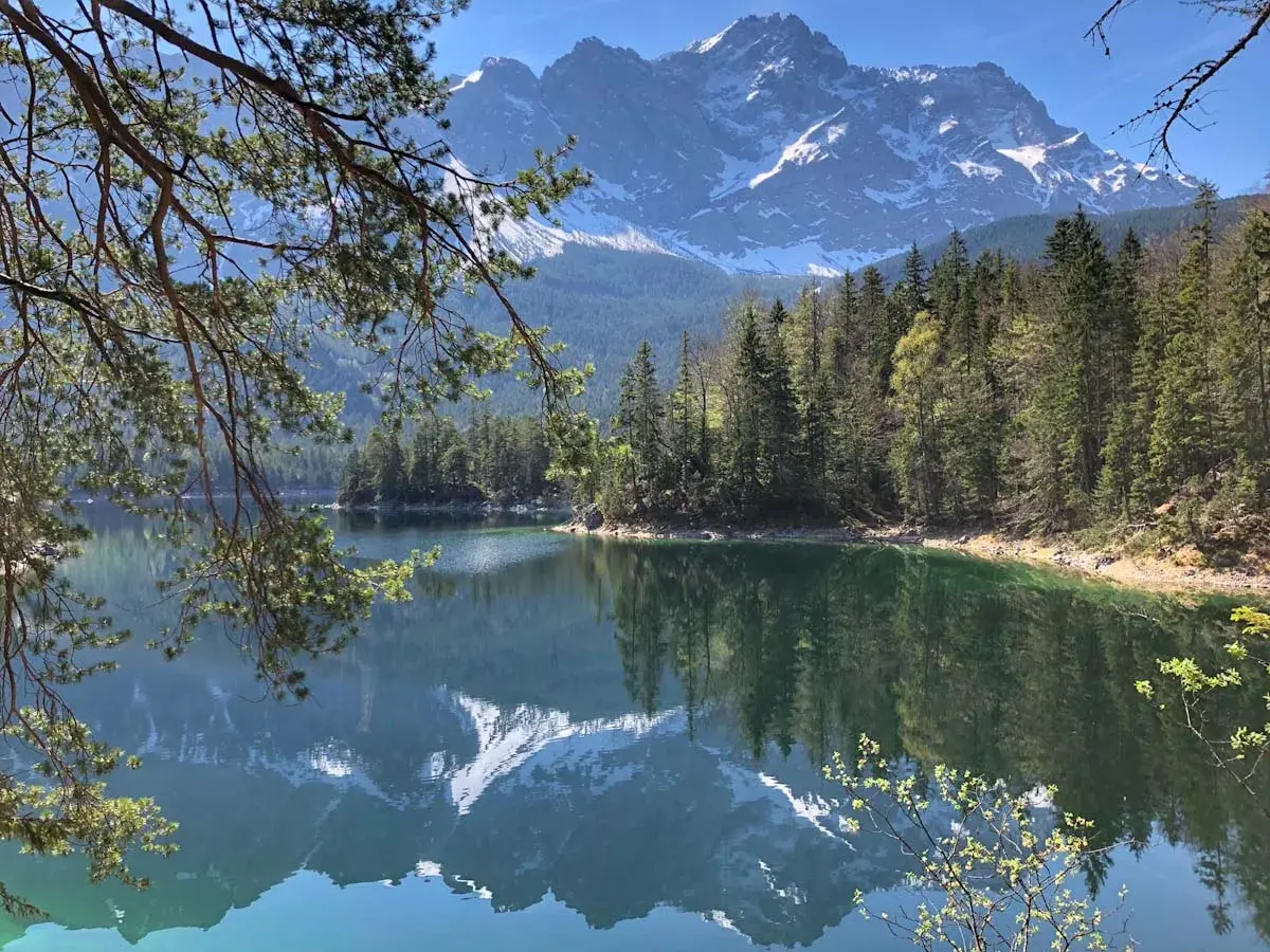 Spiegelglatter See mit Bergpanorama in den bayerischen Alpen. Nadelbäume säumen das Ufer, im Hintergrund ragen schneebedeckte Gipfel auf.