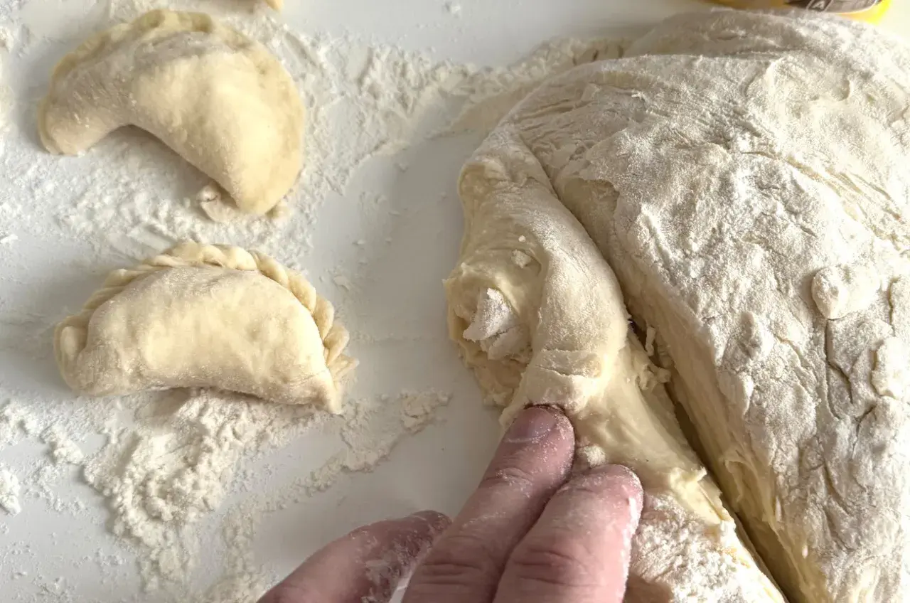 Ręce formują ciasto na pierogi, obok leżą już ulepione pierożki.