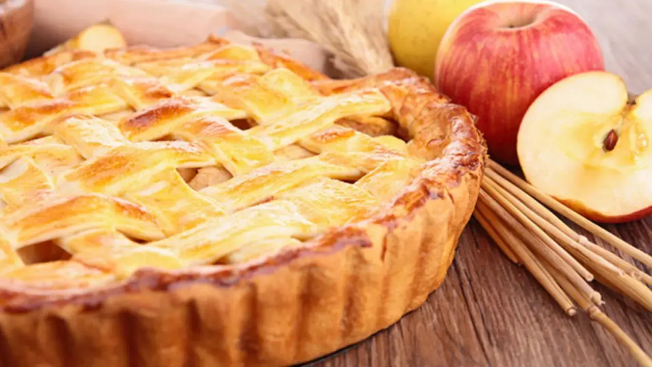 Tarta de manzana dorada con rejilla de masa, junto a manzanas frescas y palitos de canela.