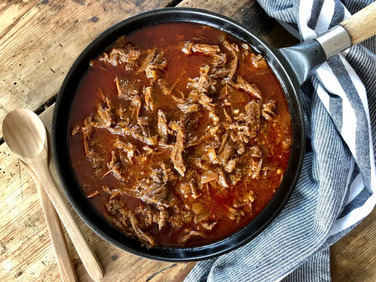 Saftigt pulled beef i en mustig sås i gjutjärnspanna, redo att serveras med träskedar bredvid.