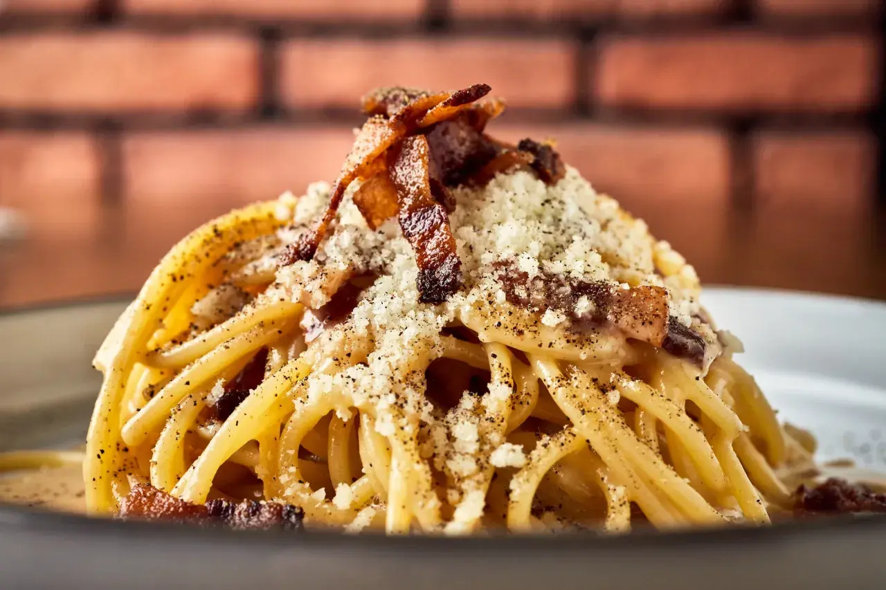 Pyszny makaron włoski carbonara, posypany parmezanem i chrupiącym boczkiem, na tle ceglanej ściany.