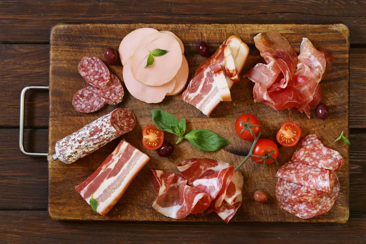 Assortiment de différents types de charcuterie : saucisson, jambon cru, bacon, mortadelle, accompagnés de tomates cerises et d'olives.
