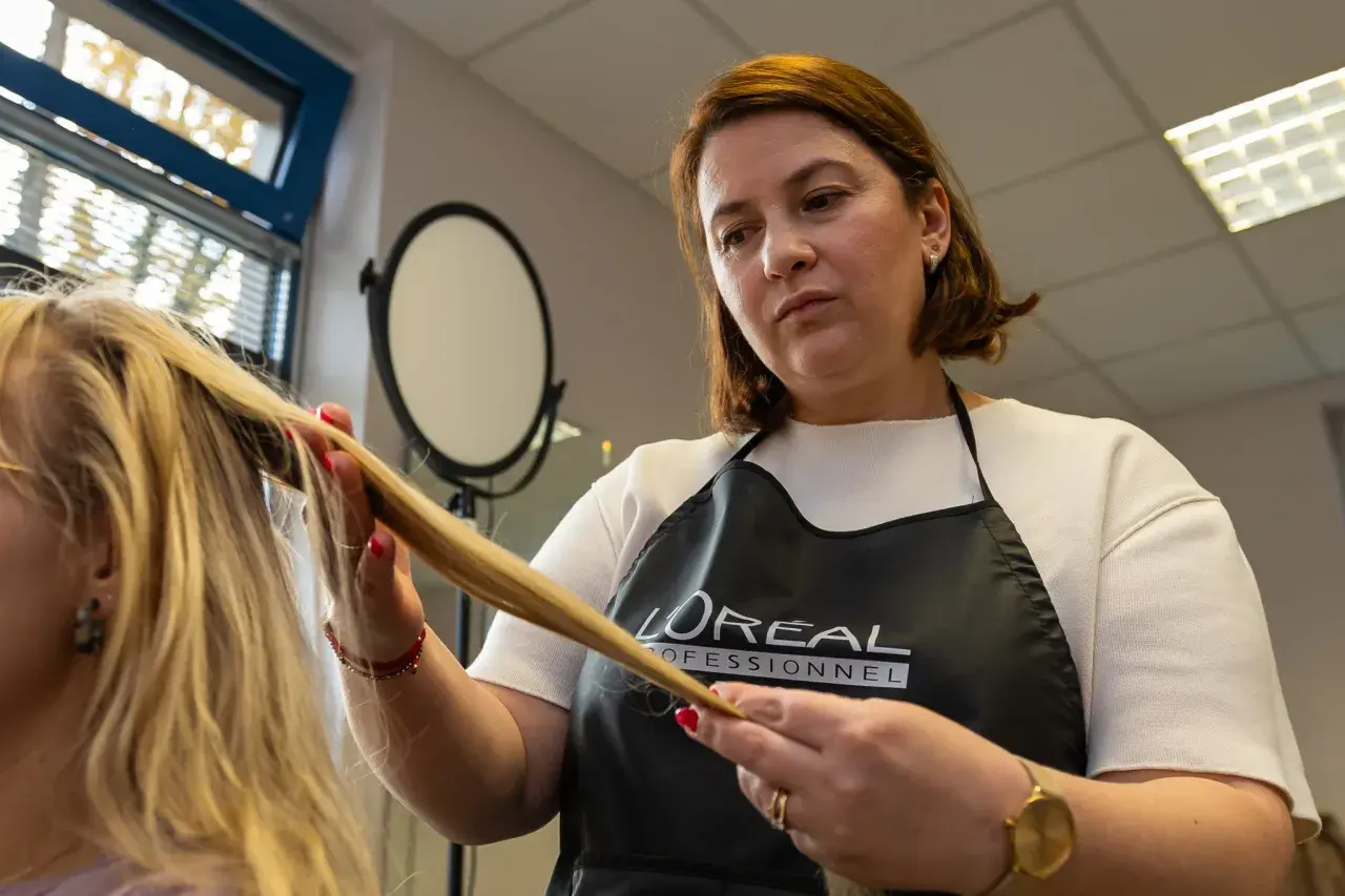 Fryzjerka w fartuchu L'Oréal Professionnel wykonuje podcinanie końcówek blond włosów.