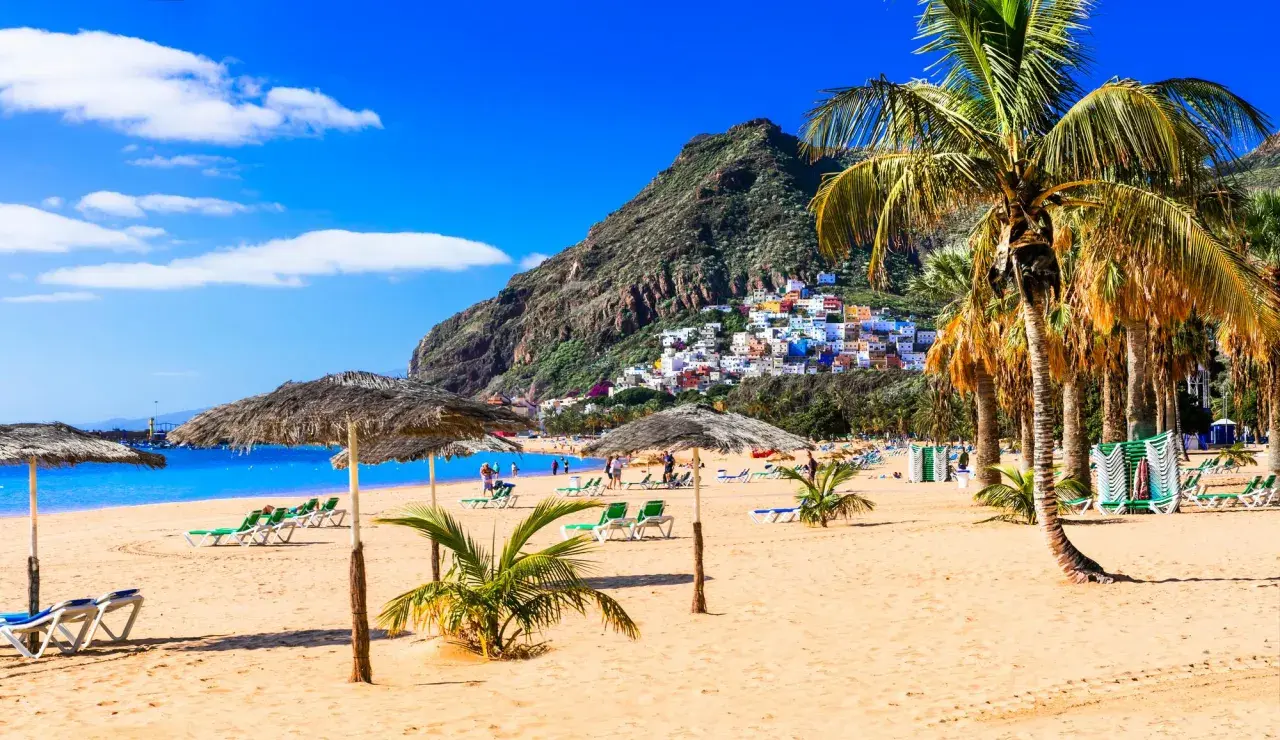Plage de sable doré, palmiers, parasols et maisons colorées perchées sur une colline. Une invitation à visiter Tenerife pour des vacances de rêve.