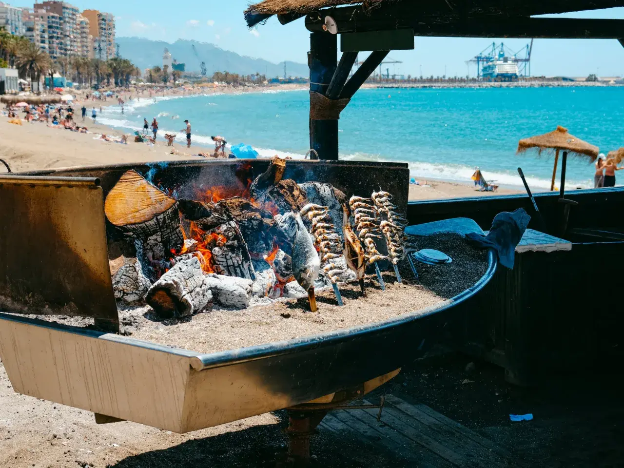 Pescado a la brasa en un chiringuito del paseo marítimo de Málaga. El mar y la playa con gente al fondo.