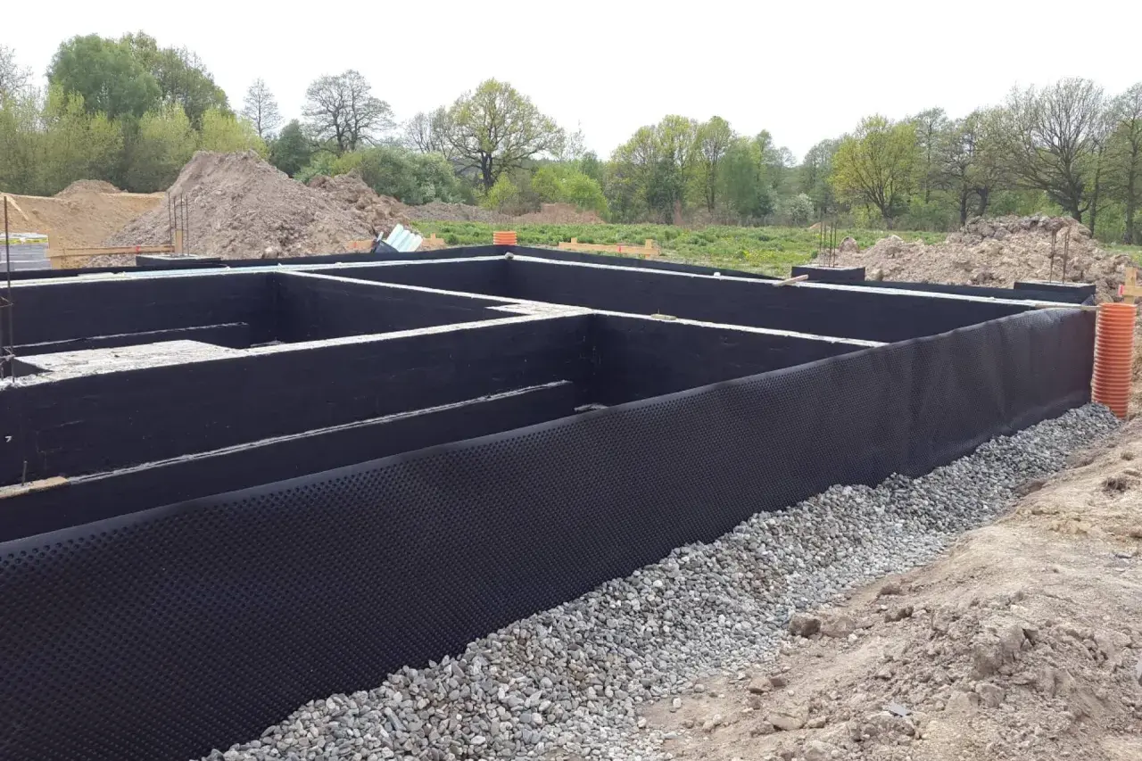 Fundament domu w budowie, z czarną izolacją i otoczakami.