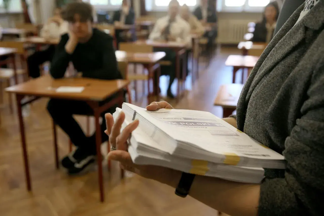 Ręka trzyma stos arkuszy egzaminacyjnych z napisem "JĘZYK POLSKI". W tle uczniowie siedzą przy ławkach.